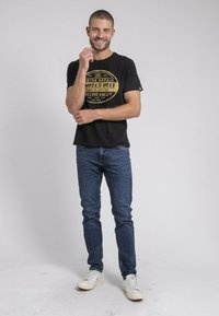 T-shirt noir avec un design graphique doré, associé à un jean en denim bleu et des baskets blanches. Le modèle se tient souriant, les mains positionnées de manière décontractée.