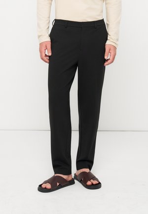 Homme portant un pantalon noir droit et des sandales marron à brides croisées, debout devant un fond blanc uni.