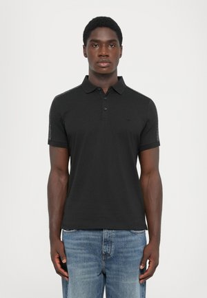 Black polo shirt with a collar, three buttons, and textured side panels; paired with light blue denim jeans. Minimal branding on chest.