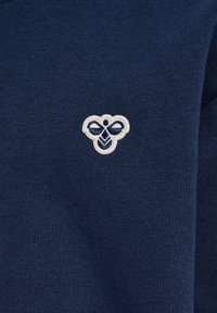 Granatowy dzianinowy materiał z małym białym haftowanym logo pszczoły umieszczonym na środku na wysokości klatki piersiowej ubrania.