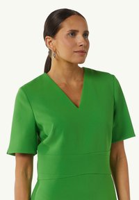 Jasnozielona sukienka z dekoltem w kształcie litery V, krótkimi rękawami i dopasowaną talią. Gładki materiał o wyraźnej sylwetce i subtelnych detalach szwowych.