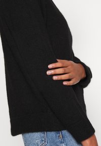 Schwarzer Strickpullover mit lockerem Schnitt, überschnittenen Schultern und gerippten Bündchen, kombiniert mit hellen Denim-Jeans. Weiche Textur sichtbar.