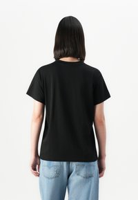 Person with shoulder-length dark hair wearing a plain black T-shirt and light blue jeans standing facing away against a white background.