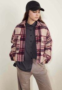 Veste en polaire à carreaux rose et bordeaux avec deux poches avant, fermeture à boutons, et une casquette noire portée sur une chemise à boutons sombre et un pantalon beige.