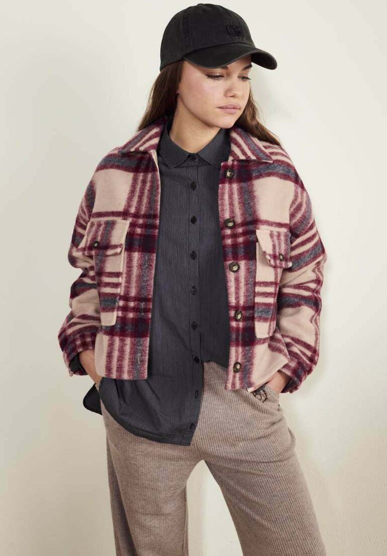 Veste en polaire à carreaux rose et bordeaux avec deux poches avant, fermeture à boutons, et une casquette noire portée sur une chemise à boutons sombre et un pantalon beige.