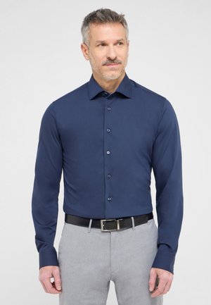 Marineblaue Langarmbluse mit glatter Textur, Knopfleiste, spitzem Kragen und kontrastierenden Knöpfen, kombiniert mit grauen Hosen und einem schwarzen Gürtel.