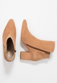 Botas de ante color marrón claro hasta el tobillo, con punta afilada y cierre de cremallera lateral. Presentan un tacón ancho y una textura suave.