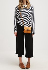 Kvinde iført sorte brede bukser, stribet langærmet skjorte, brun læder crossbody taske og brune snørebåndssko, stående foran en ensfarvet baggrund.