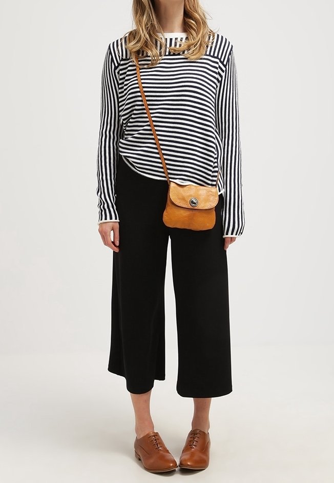 Kvinde iført sorte brede bukser, stribet langærmet skjorte, brun læder crossbody taske og brune snørebåndssko, stående foran en ensfarvet baggrund.