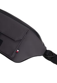 Riñonera de nylon gris con un bolsillo frontal con tapa, herrajes en negro y una pequeña etiqueta con el logo. Cuenta con una correa duradera para ajuste y comodidad.