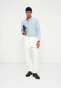 Camisa azul clara de botões com um bolso no peito, calças brancas ajustadas e sapatos loafers pretos; modelo segura uma bolsa preta. Design simples e limpo.