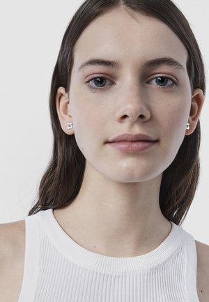 Giovane donna con capelli castani lisci indossa orecchini a bottone a forma di fiore d'argento e una canotta bianca a coste su uno sfondo semplice.