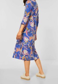 Blaues blumiges Midi-Kleid mit Dreiviertelärmeln, das mehrfarbige Blumenmuster und eine taillierte Taille aufweist. Kombiniert mit lässigen beigen Schuhen.