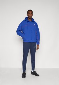 Blue nylon jacket with a hood and red accents, paired with navy athletic pants and black shoes. Features Nike logos on both garments.