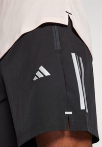Pantalones cortos deportivos negros con un bolsillo lateral con cremallera, que presentan rayas reflectantes plateadas y el logo de Adidas en el dobladillo izquierdo. Textura suave, diseño aerodinámico.