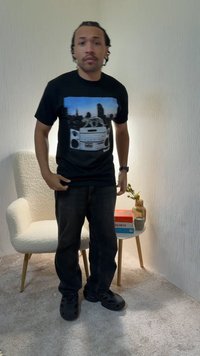 Man in black graphic t-shirt and dark pants standing next to a white textured armchair and small round table with books, vase, and cup.