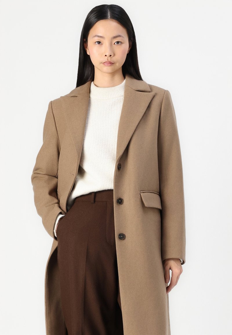 Lungo cappotto beige con colletto a risvolto, chiusura con un solo bottone e tasche laterali. Indossato sopra un dolcevita crema e pantaloni marroni.