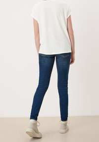 Bílý krátký rukávový top v kombinaci s tmavě modrými úzkými džíny a béžovými kotníkovými botami, zobrazující nenucený outfit zezadu.