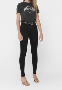 Jeans negros ajustados de cintura alta combinados con una camiseta gráfica de color carbón que presenta un diseño circular y texto. Finalizado con un cinturón negro y sandalias.