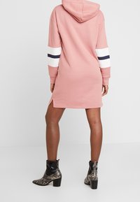 Femme portant une robe à capuche rose avec des rayures blanches et marine sur les manches, associée à des bottines à talons en serpent noir et gris, se tenant devant un fond blanc.