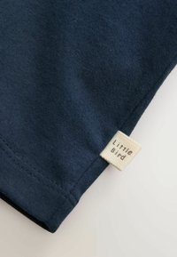 Tamsiai mėlyna medžiaga su mažu įsiūtu smėlio spalvos etiketės gabaliuku siūlėje, kur parašyta „little bird“ mažosiomis raidėmis.