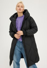 Manteau doudoune noir avec capuche, présentant une texture matelassée et une fermeture éclair à l'avant. Porté sur un pull en maille torsadée violet et un jean bleu.