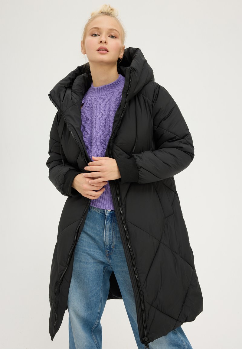 Manteau doudoune noir avec capuche, présentant une texture matelassée et une fermeture éclair à l'avant. Porté sur un pull en maille torsadée violet et un jean bleu.