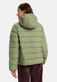 Chaqueta verde acolchada con capucha, que presenta una textura acolchada y costuras horizontales. Combinada con pantalones de pana marrones.