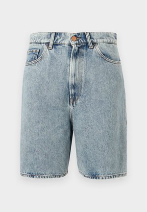 Mujer vestida con top cruzado y shorts de mezclilla azul claro, sosteniendo un bolso de mano azul marino y usando botas negras de tacón hasta la rodilla, de pie contra un fondo blanco.