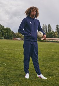 Marineblå treningsdrakt med hette og lommer, hvite og rosa fotballsko, holder en grå fotball på en gressbane med trær i bakgrunnen.