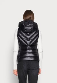 Zwarte gewatteerde bodywarmer met capuchon, voorzien van chevron-stiksels en een glanzende afwerking, gecombineerd met een wit langermig shirt en donkere jeans.