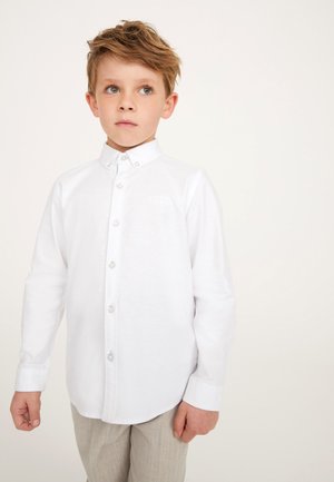 Ragazzo con capelli castano chiaro che indossa una camicia bianca abbottonata a maniche lunghe e pantaloni beige, in piedi davanti a uno sfondo chiaro e uniforme.