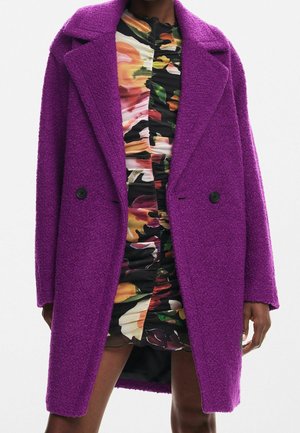 Femme portant un manteau violet texturé à double boutonnage sur une mini-robe fleurie froncée aux nuances de noir, orange, vert et rose.