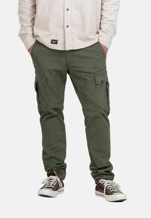 Mann trägt olivgrüne Cargohosen mit Klapptaschen, beige Hemd mit Knöpfen und braune Low-Top-Sneaker mit weißen Schnürsenkeln, Hände in den Taschen.