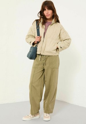 Beige gesteppte Jacke mit hohem Kragen, olivgrüne Cargo-Hosen und hellen Sneakers; trägt eine kleine blaue Umhängetasche.