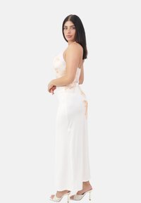 Femme aux longs cheveux noirs portant une robe maxi sans manches blanche avec des motifs floraux de pêches claires et des sandales blanches à talons hauts, se tenant de profil.