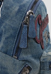 Mochila de mezclilla con acabado azul, que presenta un logo burdeos, herrajes plateados, tiradores de cremallera texturizados y un sutil desgaste.