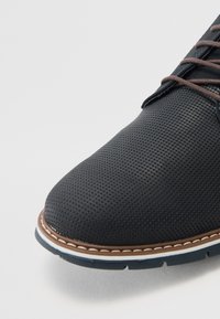 Zapato negro perforado con punta redonda, cordones marrón oscuro y un contraste de costura marrón claro a lo largo de la suela. Diseño minimalista.