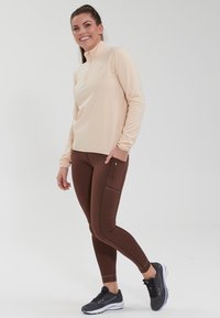 Lyse ferskenfarvet quarter-zip langærmet top parret med brune tætsiddende leggings med en sidelomme med lynlås, båret med sorte sneakers.