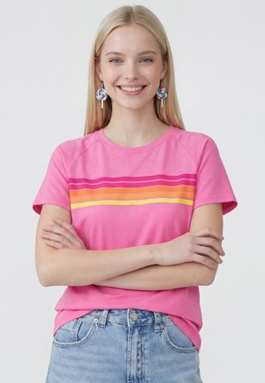 Jonge vrouw met lang blond haar glimlacht, draagt een roze T-shirt met kleurrijke strepen en oorbellen in de vorm van regenbooglollies, armen gekruist.
