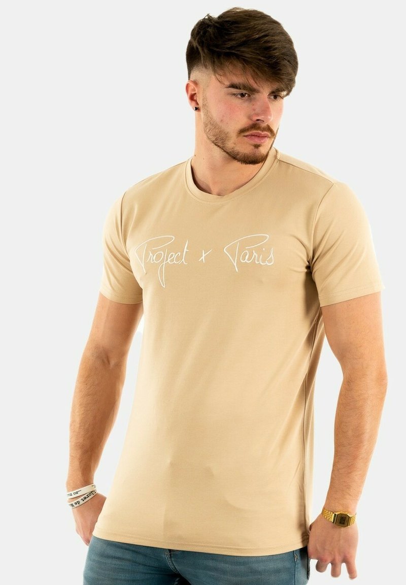 Camiseta de manga corta beige con texto en cursiva blanca "Project x Paris". Fabricada en tejido suave, presenta un diseño clásico de cuello redondo.