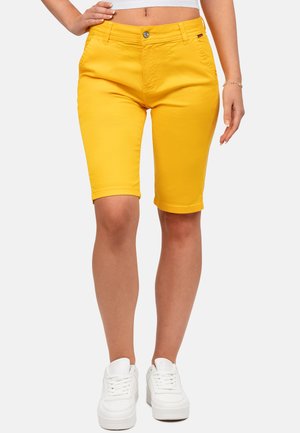 Mujer con pantalones cortos amarillos hasta la rodilla, top blanco corto y zapatillas blancas con plataforma, de pie con una mano en la cadera.