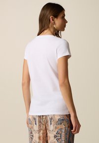 Mujer con cabello largo y castaño que lleva una camiseta blanca sencilla de manga corta y pantalones beige, azul y naranja con estampado, de pie de perfil mirando hacia la derecha.