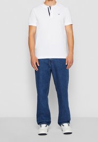 Vit piké med treknappslås och marinblå-röd detalj. Blå denimjeans. Vita sneakers. Avslappnad passform, korta ärmar, ren design.