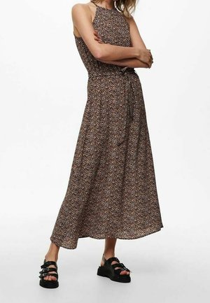 Femme portant une robe maxi sans manches marron à motif floral avec une ceinture nouée à la taille et des sandales à plateforme noires, debout les bras croisés.