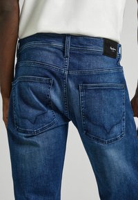 Jeans de mezclilla azul oscuro con corte recto, que presentan un parche de logo visible, dos bolsillos traseros con un patrón de costura distintivo.