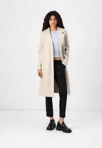 Lys beige ullkåpe med spiss krage, åpen front og lange ermer, lagd over en lys blå cropped teksturert topp og svarte bukser.