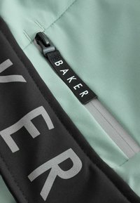 Svetlozelená tkanina s čiernym panelom s výrazným textom. Na boku je viditeľný zips s visačkou označenou "BAKER".