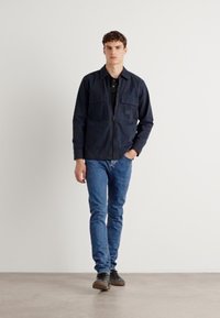 Chaqueta de cremallera de un azul marino oscuro con dos bolsillos en el pecho, combinada con unos vaqueros azules y zapatos oscuros. El modelo está de pie frente a un fondo blanco liso.