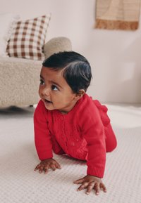 Rode gebreide trui met bloemmotieven, ronde halslijn en ruches aan de zoom; kruipende baby op een crèmekleurig gestructureerd kleed; op de achtergrond beige meubels.